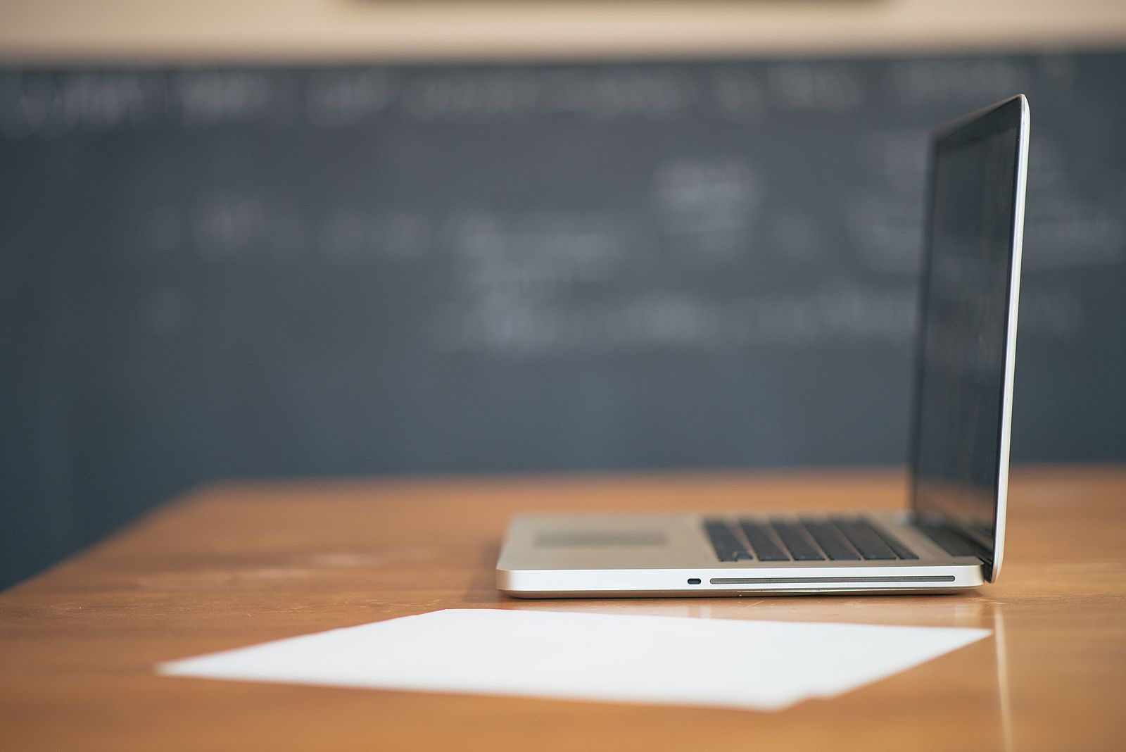 A laptop on a desk next to a piece of paper and chalkboard in the background