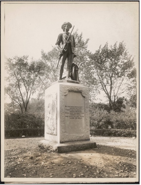 Black and white photo of a statue depicting a minuteman in a historical uniform holding a rifle. The statue stands on a stone pedestal with an inscription. The background features trees and a park-like setting.