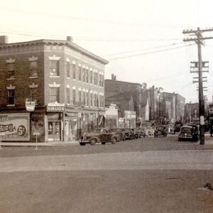 Black and white photo of Oak Street in New Haven, Connecticut. A corner drugstore with a vintage sign is prominent. Classic cars are parked along the street.