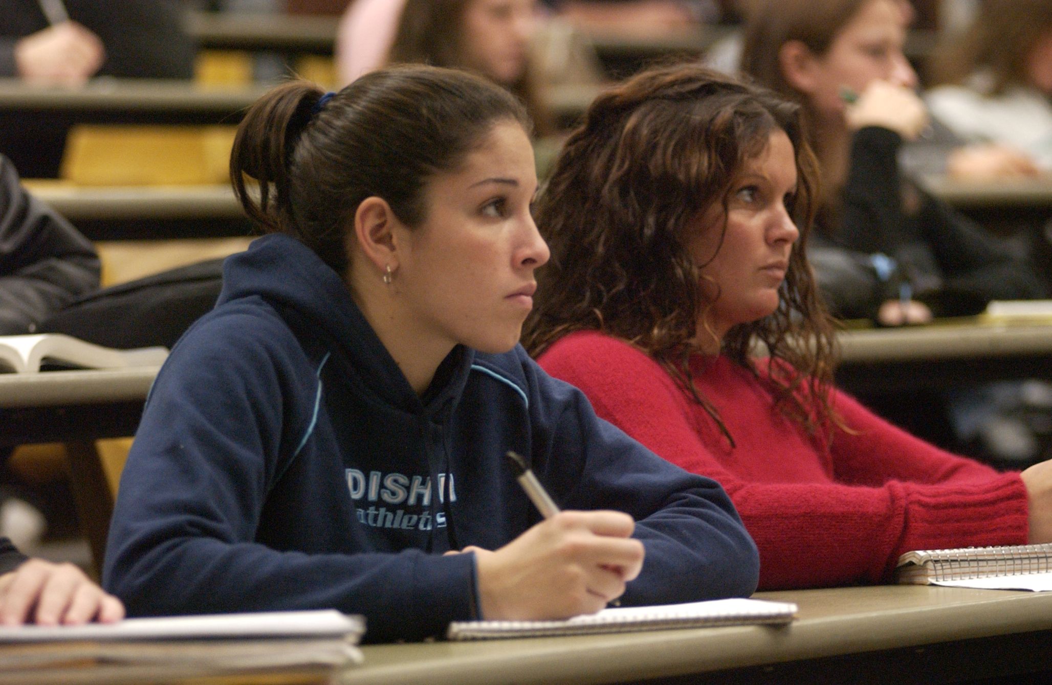 Two female students in a lecture hall listening and taking notes.