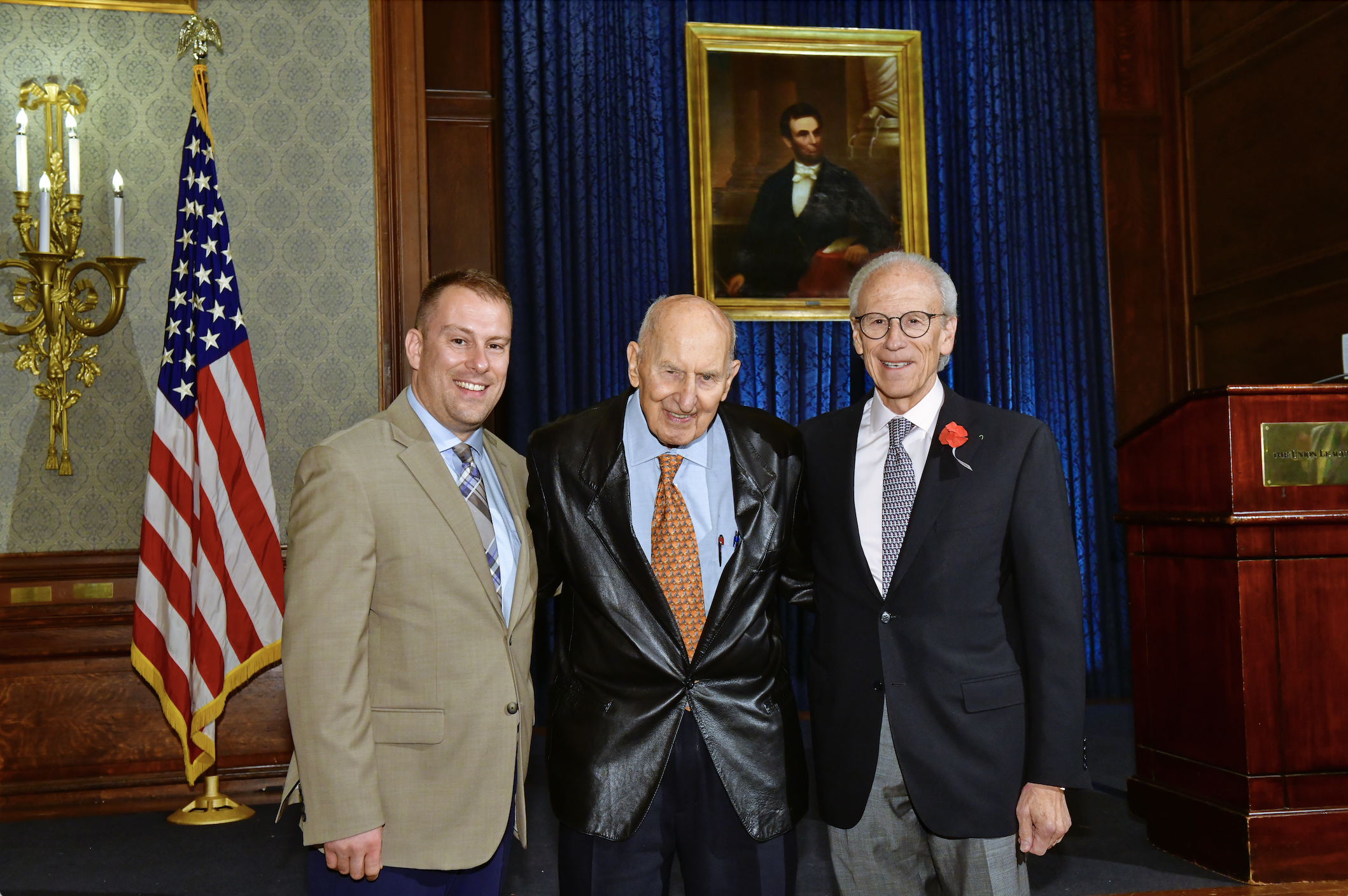 JMC President Hans Zeiger, Chairman Emeritus and Founder Jack Miller, and Chairman Michael Weiser with an American flag and Abraham Lincoln portrait