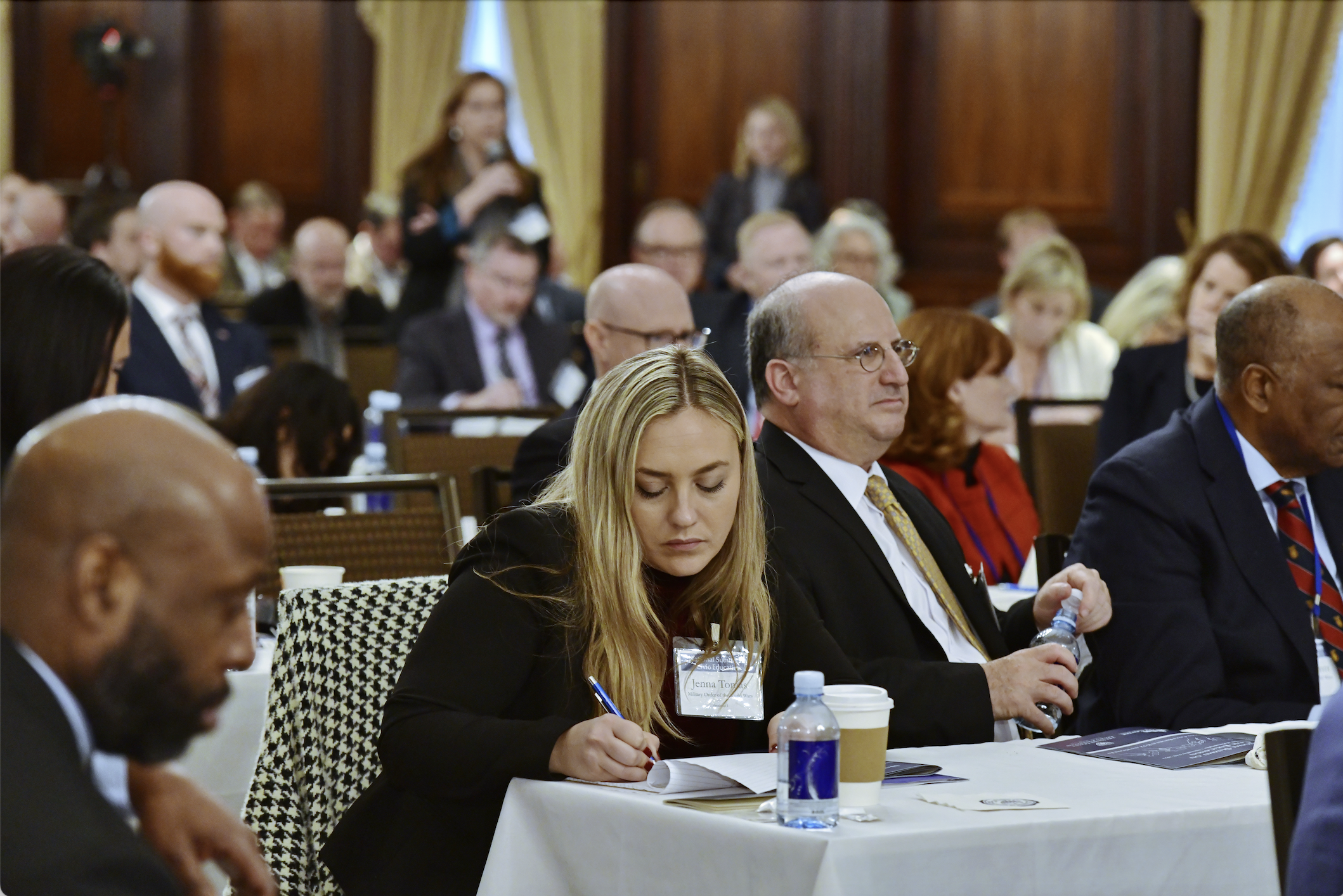 Female audience member writing notes with a participant speaking in the background at JMC's National Summit on Civic Education.